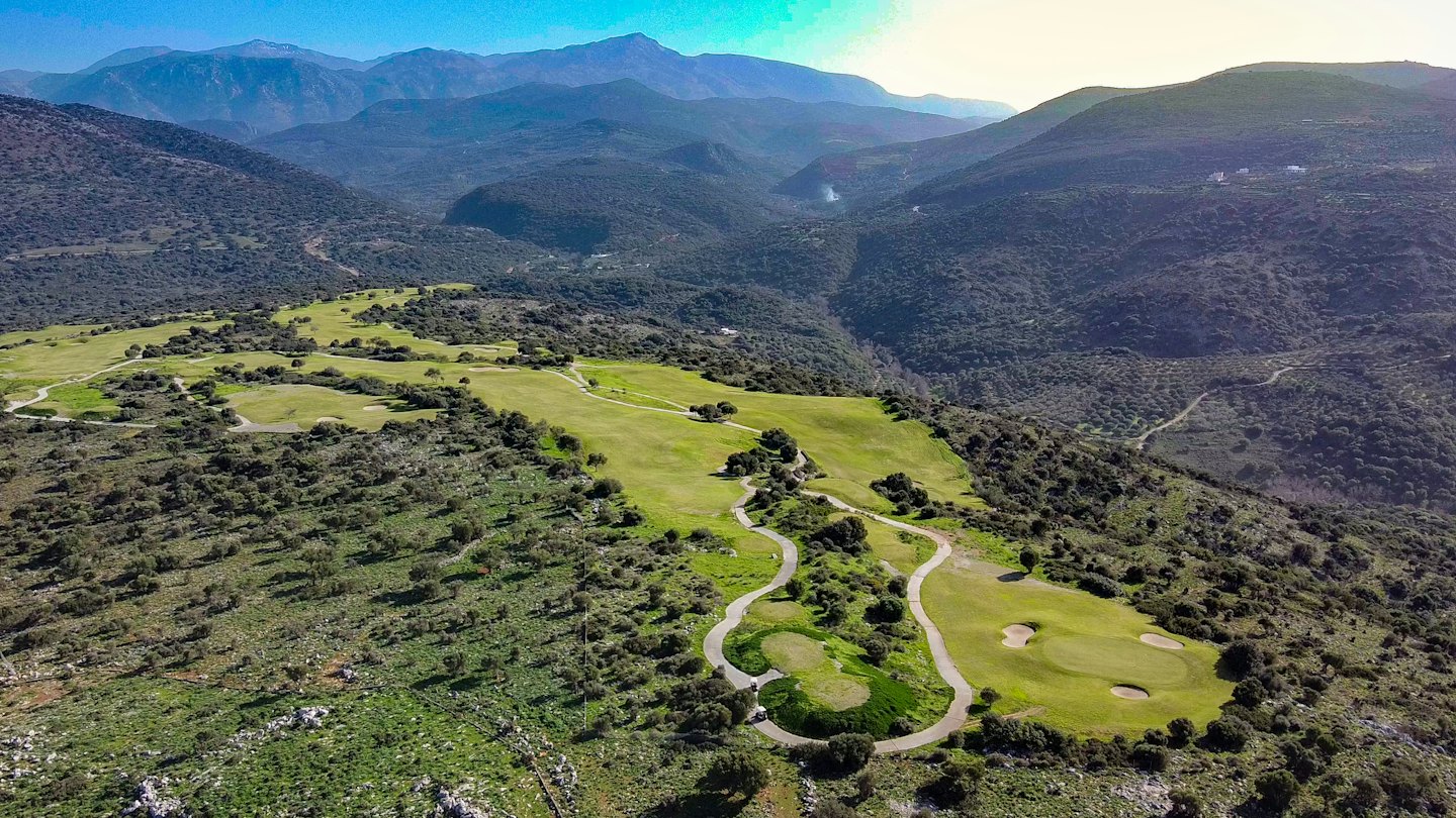 Golf course winding through a green valley with scrub-covered mountains.
